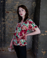 This vintage 1980s wrap blouse, crafted from luxurious pure silk, features a vibrant all-over rose print in rich red, pink, and green hues. The draped silhouette flows effortlessly, while a sculptural rose detail on the right side adds a bold, romantic twist. No tie needed – just slip it on and let the look speak for itself!