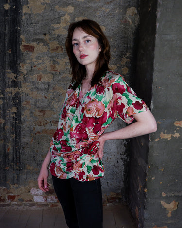 This vintage 1980s wrap blouse, crafted from luxurious pure silk, features a vibrant all-over rose print in rich red, pink, and green hues. The draped silhouette flows effortlessly, while a sculptural rose detail on the right side adds a bold, romantic twist. No tie needed – just slip it on and let the look speak for itself!