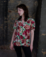 This vintage 1980s wrap blouse, crafted from luxurious pure silk, features a vibrant all-over rose print in rich red, pink, and green hues. The draped silhouette flows effortlessly, while a sculptural rose detail on the right side adds a bold, romantic twist. No tie needed – just slip it on and let the look speak for itself!