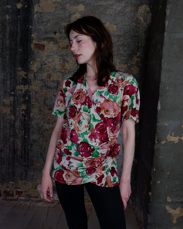This vintage 1980s wrap blouse, crafted from luxurious pure silk, features a vibrant all-over rose print in rich red, pink, and green hues. The draped silhouette flows effortlessly, while a sculptural rose detail on the right side adds a bold, romantic twist. No tie needed – just slip it on and let the look speak for itself!