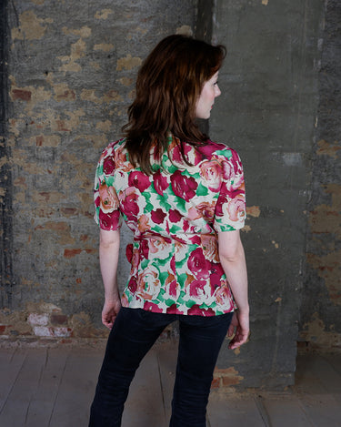 This vintage 1980s wrap blouse, crafted from luxurious pure silk, features a vibrant all-over rose print in rich red, pink, and green hues. The draped silhouette flows effortlessly, while a sculptural rose detail on the right side adds a bold, romantic twist. No tie needed – just slip it on and let the look speak for itself!