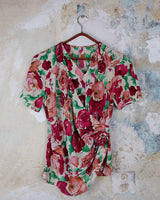 This vintage 1980s wrap blouse, crafted from luxurious pure silk, features a vibrant all-over rose print in rich red, pink, and green hues. The draped silhouette flows effortlessly, while a sculptural rose detail on the right side adds a bold, romantic twist. No tie needed – just slip it on and let the look speak for itself!