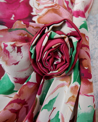 This vintage 1980s wrap blouse, crafted from luxurious pure silk, features a vibrant all-over rose print in rich red, pink, and green hues. The draped silhouette flows effortlessly, while a sculptural rose detail on the right side adds a bold, romantic twist. No tie needed – just slip it on and let the look speak for itself!
