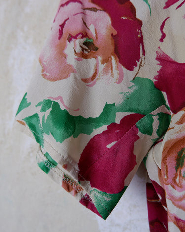 This vintage 1980s wrap blouse, crafted from luxurious pure silk, features a vibrant all-over rose print in rich red, pink, and green hues. The draped silhouette flows effortlessly, while a sculptural rose detail on the right side adds a bold, romantic twist. No tie needed – just slip it on and let the look speak for itself!