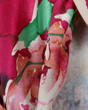 This vintage 1980s wrap blouse, crafted from luxurious pure silk, features a vibrant all-over rose print in rich red, pink, and green hues. The draped silhouette flows effortlessly, while a sculptural rose detail on the right side adds a bold, romantic twist. No tie needed – just slip it on and let the look speak for itself!