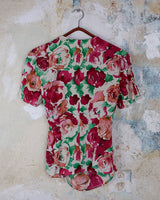 This vintage 1980s wrap blouse, crafted from luxurious pure silk, features a vibrant all-over rose print in rich red, pink, and green hues. The draped silhouette flows effortlessly, while a sculptural rose detail on the right side adds a bold, romantic twist. No tie needed – just slip it on and let the look speak for itself!