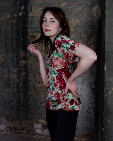 This vintage 1980s wrap blouse, crafted from luxurious pure silk, features a vibrant all-over rose print in rich red, pink, and green hues. The draped silhouette flows effortlessly, while a sculptural rose detail on the right side adds a bold, romantic twist. No tie needed – just slip it on and let the look speak for itself!