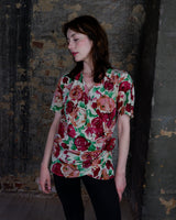This vintage 1980s wrap blouse, crafted from luxurious pure silk, features a vibrant all-over rose print in rich red, pink, and green hues. The draped silhouette flows effortlessly, while a sculptural rose detail on the right side adds a bold, romantic twist. No tie needed – just slip it on and let the look speak for itself!