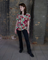This vintage 1980s wrap blouse, crafted from luxurious pure silk, features a vibrant all-over rose print in rich red, pink, and green hues. The draped silhouette flows effortlessly, while a sculptural rose detail on the right side adds a bold, romantic twist. No tie needed – just slip it on and let the look speak for itself!