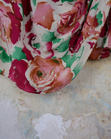This vintage 1980s wrap blouse, crafted from luxurious pure silk, features a vibrant all-over rose print in rich red, pink, and green hues. The draped silhouette flows effortlessly, while a sculptural rose detail on the right side adds a bold, romantic twist. No tie needed – just slip it on and let the look speak for itself!