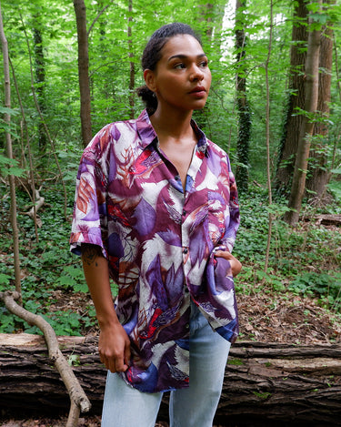 This 1990s Thai silk shirt with its chest pocket showcases a vibrant bird-and-leaf pattern in rich purples, reds, and blues. The luxurious silk fabric offers a smooth, lightweight feel with an eye-catching, exotic flair. Vintage, loud, and anything but boring.